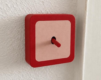 May include: A square light switch mounted on a white wall, featuring a bold red outer frame and a light pink central plate. The switch mechanism consists of a single, thin, red cylindrical toggle lever protruding from the center.