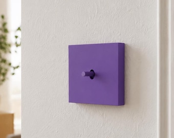 May include: A square, matte purple light switch with a central toggle lever mounted on a white wall. The switch features a flat, solid-colored faceplate and a matching cylindrical toggle that protrudes from the center of the square plate.