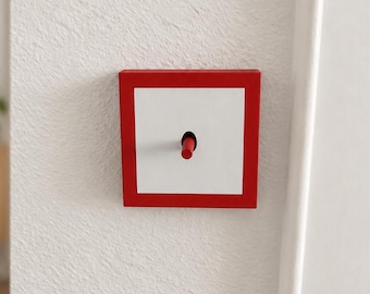 May include: A square light switch mounted on a wall, featuring a white center plate framed by a bold red border. A small, cylindrical red toggle lever protrudes from the center of the white plate, allowing for manual operation of the electrical switch.