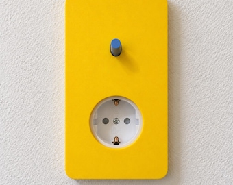 May include: A rectangular yellow wall plate featuring a single blue toggle switch positioned above a circular white electrical outlet. The plate has rounded corners and a smooth, flat surface, providing a contrast between the yellow face and the white socket.