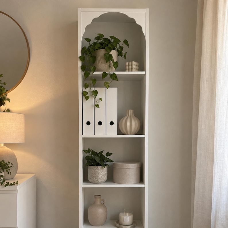 May include: A tall white wooden bookshelf featuring an arched top design with four shelves. The shelves are decorated with various items including a trailing green plant in a pot, three white binders, a ribbed vase, a round box, and candles.