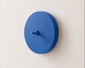 May include: A circular blue wall hook with a matte finish. The object consists of a flat, round base plate with a small, cylindrical peg extending horizontally from the center, designed for mounting items on a vertical surface.
