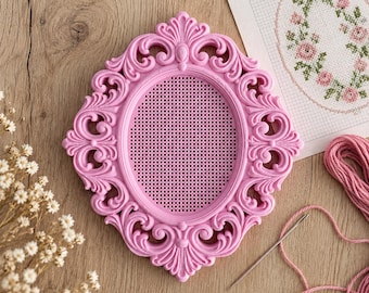 May include: An oval pink frame with an ornate, scrolled border and a central mesh grid. Beside it lies a piece of white fabric featuring a cross-stitch pattern of pink roses and green leaves, along with a silver needle and pink embroidery thread.