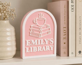 May include: An arched pink and white sign featuring a raised white graphic of a stack of three books topped with a steaming coffee cup. The text EMILY'S LIBRARY is embossed in white capital letters below the books on the pink background.