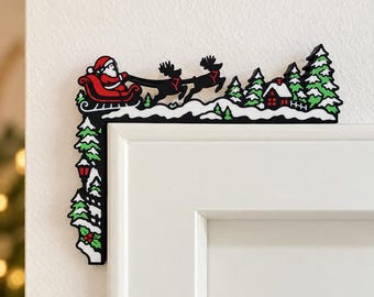 May include: A black, white, red, and green L-shaped door corner sign featuring a silhouette of Santa Claus in a sleigh pulled by two reindeer, flying over a snowy landscape with pine trees, a small house, and a street lamp.