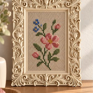 Könnte beinhalten: Ein rechteckiger cremefarbener Rahmen mit kunstvollen Schnitzereien umrandet eine perforierte Platte mit einem Kreuzstich-Blumenmuster. Die Stickerei zeigt eine große rosa Blume, kleinere rosa Knospen, grüne Blätter und kleine blaue Blüten.