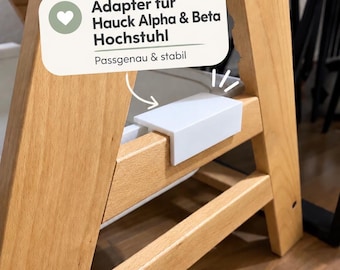 May include: A white plastic adapter attached to a light-colored wooden high chair frame. A text overlay reads: Adapter für Hauck Alpha & Beta Hochstuhl, Passgenau & stabil. The adapter is rectangular and fits over the wooden crossbar.