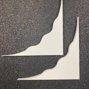 May include: Two white shelf brackets with a smooth finish and a distinct wavy, curved edge design. Each bracket features a right-angle corner structure, providing a clean and simple profile for mounting purposes. The surfaces are matte and solid white.