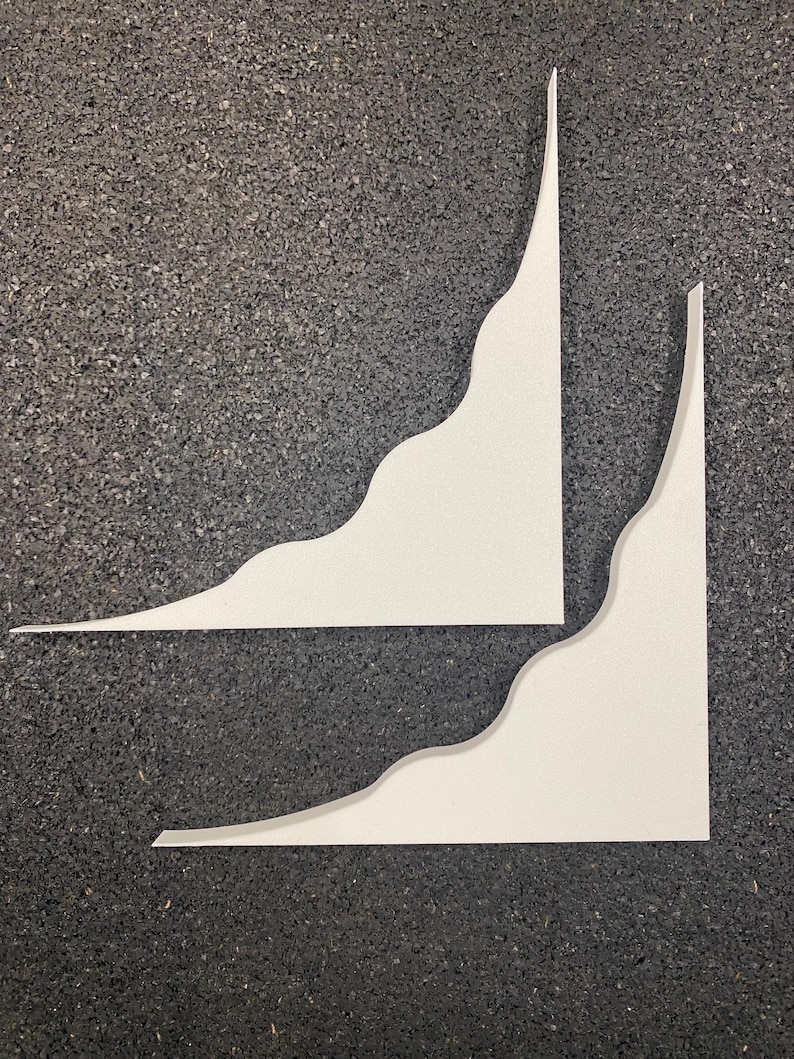 May include: Two white shelf brackets with a smooth finish and a distinct wavy, curved edge design. Each bracket features a right-angle corner structure, providing a clean and simple profile for mounting purposes. The surfaces are matte and solid white.