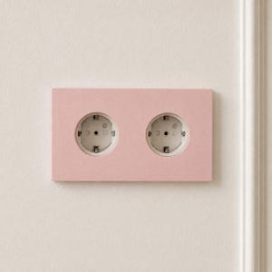 Può includere: Una placca rettangolare per doppia presa elettrica a muro con una finitura in tinta unita rosa chiaro opaco. La placca presenta due prese elettriche bianche circolari con contatti di messa a terra standard montate orizzontalmente.