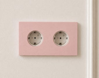 May include: A rectangular double electrical wall outlet featuring a matte light pink faceplate. The unit contains two circular white sockets with standard grounding contacts, mounted flush against a plain off-white wall surface.