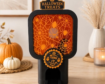 May include: A black and orange Halloween treats dispenser featuring a square frame with a spiderweb and spider cutout design. The top sign reads HALLOWEEN TREATS and a circular front dial features the text TURN IT with ghost and pumpkin icons.