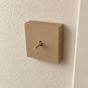 May include: A square light switch with a matte beige finish and a central toggle lever. The switch plate features a distinct ribbed texture along its top and side edges, providing a structured appearance against the flat wall surface.