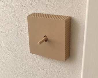 May include: A square light switch cover in a solid tan color with a ribbed texture along the outer edges. The cover features a central circular opening with a matching tan toggle switch lever extending outward from the center of the plate.