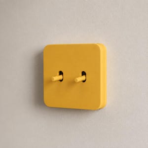 May include: A square, mustard yellow light switch plate featuring two matching toggle switches. The plate has rounded corners and a smooth, matte finish, with the two cylindrical toggles protruding from vertical slots in the center of the unit.