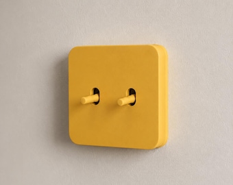 May include: A square, mustard yellow light switch plate featuring two toggle switches. The plate has rounded corners and a smooth, matte finish, with the two matching yellow toggles positioned horizontally in the center of the faceplate.
