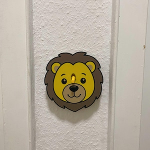 Peut inclure: Une plaque de recouvrement d'interrupteur en forme de tête de lion. Le lion a un visage jaune, une crinière brune et des contours noirs. Un petit interrupteur à bascule jaune dépasse du centre du visage du lion, entre les yeux et le nez.