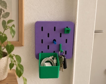 May include: A square purple pegboard mounted on a wall, featuring a green storage bin, a blue peg, a green hook holding a set of keys, and a small green block-style attachment. The green bin contains a white charging cable and a power adapter.