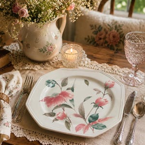 May include: An octagonal white ceramic dinner plate featuring a painted pink rose and green leaf pattern. The table setting includes a floral napkin with a woven ring, silver-toned flatware, a pink textured glass goblet, and a clear glass tumbler.