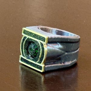 May include: A metallic ring featuring a rectangular face with a dark green textured inlay and a central circular green accent. The band has a dark grey finish with a layered, ridged design on the sides and a gold-toned border around the face.