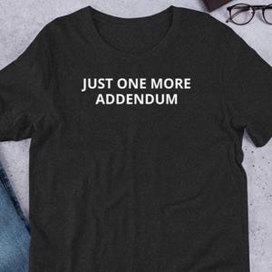 Può includere: T-shirt grigio scuro con la scritta bianca "JUST ONE MORE ADDENDUM". La maglietta a maniche corte è realizzata in materiale morbido. La maglietta è piegata su una superficie grigio chiaro.
