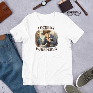 Puede incluir: Camiseta blanca con un dibujo de un hombre con traje y sombrero de vaquero susurrando a una caja fuerte. El texto "LOCKBOX WHISPERER" está encima y debajo de la ilustración. La camiseta se muestra sobre una superficie clara.