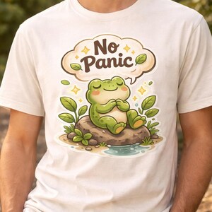 Könnte beinhalten: Weißes T-Shirt mit einem Cartoon-Frosch, der auf einem Felsen sitzt und die Worte "No Panic" in einer Sprechblase trägt. Der grüne Frosch hat die Augen geschlossen, das Design umfasst Blätter und Wasser.