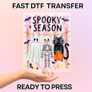 Può includere: Un foglio di trasferimento DTF rettangolare con il testo SPOOKY SEASON in lettere nere. Il disegno mostra un fantasma bianco, uno scheletro e un gatto nero che tiene una zucca arancione, appesi a un filo per il bucato con mollette di legno.