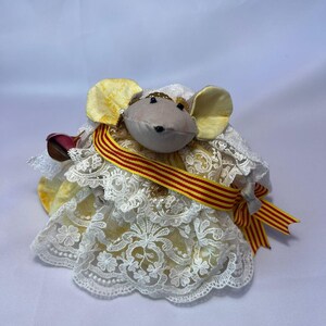 May include: A plush mouse doll wearing a yellow and white lace dress with a red and yellow striped ribbon. The mouse has large yellow ears and a gray face. The doll is a handmade craft.