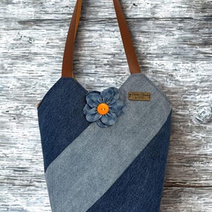 May include: Denim tote bag with brown leather handles. The bag features a diagonal stripe design in shades of blue denim and a denim flower with an orange center. A small, rectangular tag is attached to the bag.