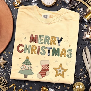 Może przedstawiać: Bladożółty t-shirt z okrągłym dekoltem z napisem MERRY CHRISTMAS w wielokolorowe wzory w kratę i paski. Poniżej tekstu znajdują się ilustracje choinki, skarpety świątecznej i gwiazdy, wszystkie z motywami w stylu patchworkowym.