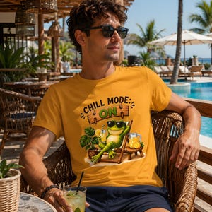 May include: A mustard yellow t-shirt featuring a graphic of a green frog wearing sunglasses, lounging in a beach chair with a drink and a pitcher of orange juice, with the text CHILL MODE: ON printed above the frog in a wooden sign style.
