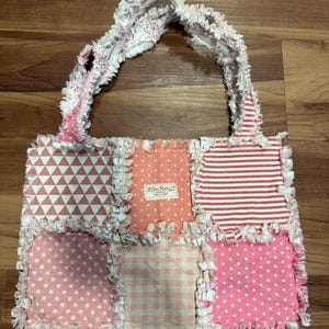May include: A patchwork tote bag with a variety of pink and white patterned fabrics. The bag features triangles, polka dots, stripes, and gingham. The edges are frayed, and the handles are made of a white floral fabric.