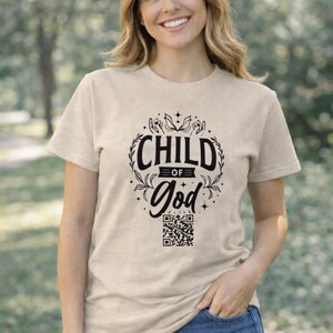 May include: Beige t-shirt with the text "CHILD OF GOD" in a black, decorative font. The design includes a floral wreath and a QR code at the bottom. The person is wearing blue jeans.