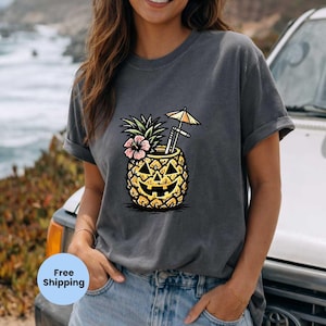 May include: Dark gray t-shirt featuring a pineapple design with a jack-o'-lantern face, a pink flower, and a small umbrella. The pineapple has a straw and a cocktail umbrella. The shirt is a casual style.
