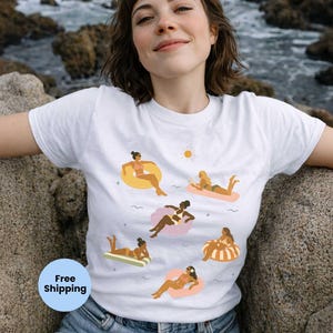 May include: White t-shirt featuring a graphic of women on inflatable floats and sunbathing. The design includes a yellow sun and various float colors. The shirt is worn by a person outdoors.