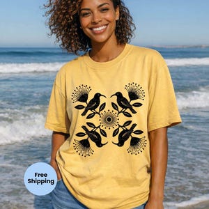 May include: A mustard yellow short-sleeve t-shirt featuring a symmetrical black graphic print of four birds and four spiky floral elements arranged in a square pattern around a central circular sun-like motif on the front of the garment.