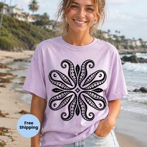 May include: A light purple short-sleeved t-shirt featuring a large, symmetrical black graphic print on the chest. The design consists of four curved, tentacle-like shapes arranged in a floral pattern, decorated with small white dots and circles.