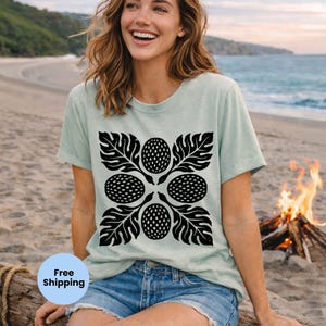 May include: A light green t-shirt featuring a black floral design with four circular elements and leaf accents. The shirt is paired with denim shorts. The image is taken on a beach with a bonfire in the background.