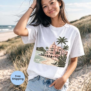 May include: White t-shirt featuring a colorful illustration of a tropical resort with palm trees, a beach, and ocean waves. The graphic is in shades of pink, green, and beige. The shirt is worn by a person in blue jeans.