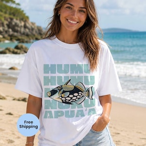 May include: White t-shirt featuring a Humuhumunukunukuāpuaʻa fish design in black, yellow, and white, with text in light blue. The shirt is worn on a beach with a blue ocean and sky in the background.