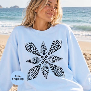 Puede incluir: Una sudadera de cuello redondo en color azul claro con un estampado gráfico central de ocho conchas marinas negras dispuestas en forma de estrella. Las conchas tienen texturas y puntos variados, creando un diseño simétrico en la prenda.