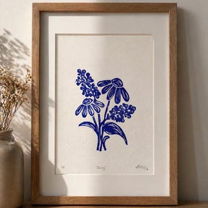 Puede incluir: Una lámina artística enmarcada con un grabado en tinta azul de un ramo floral con dos flores tipo margarita y dos racimos de flores pequeñas. La impresión está sobre papel blanco roto, titulada "Daisy" a lápiz y en un marco de madera clara.
