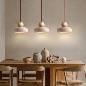 Könnte beinhalten: Drei Pendelleuchten mit hellrosa, gesprenkeltem Schirm und einer Holzkugel darüber. Die Leuchten hängen über einem Esstisch aus Holz mit Stühlen. Der Raum hat eine neutrale Farbpalette.