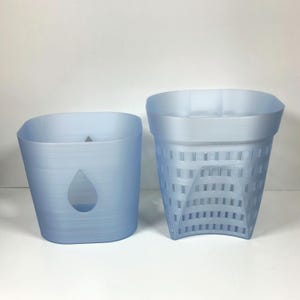 Puede incluir: Dos macetas de plástico azul claro. Una tiene forma cuadrada con un recorte de lágrima. La otra tiene un diseño de tejido de cesta. Ambas están vacías y se encuentran sobre una superficie blanca.