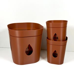 Könnte beinhalten: Ein Set aus vier braunen Pflanztöpfen aus Kunststoff. Ein großer Topf und drei kleinere Töpfe sind zu sehen. Jeder Topf hat einen tropfenförmigen Ausschnitt auf der Vorderseite. Die Töpfe sind leer und bereit zum Bepflanzen.