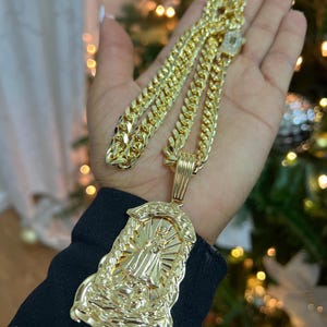 Peut inclure: Un collier doré avec un grand pendentif représentant une figure religieuse. La chaîne est de style gourmette épaisse. Le pendentif présente des détails complexes et est très réfléchissant. Le collier est présenté sur un fond flou.