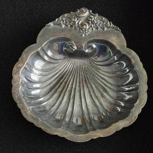 May include: A silver-colored, shell-shaped dish with a scalloped edge. The dish features a detailed, fan-like pattern radiating from the center. The top edge has an ornate, floral design. The dish is likely made of metal and is displayed against a dark background.