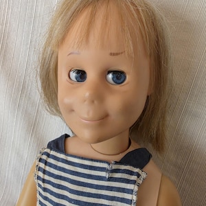 May include: A vintage doll with blonde hair and blue eyes. The doll is wearing a blue and white striped top. The doll has a smiling expression and is made of plastic.