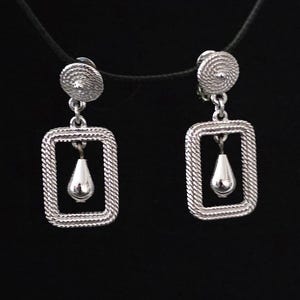 May include: A pair of silver-toned earrings. Each earring features a circular top, a rectangular frame with a rope-like texture, and a teardrop-shaped pendant. The earrings hang from a black cord against a black background.